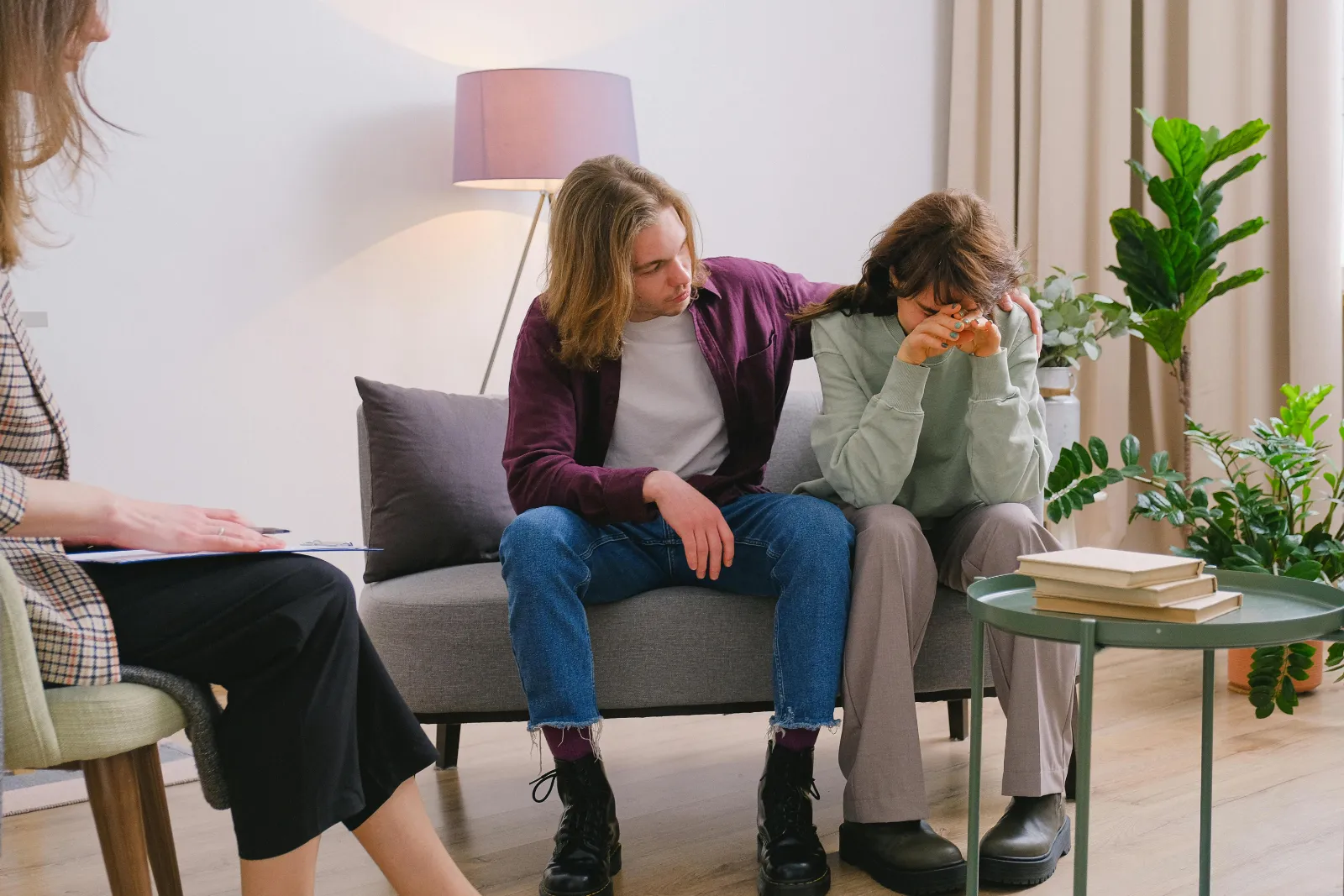 A young man with his arm around a woman sitting on a couch. They seem sad due to experiencing grief and loss of some kind and seem to be in a therapist's office