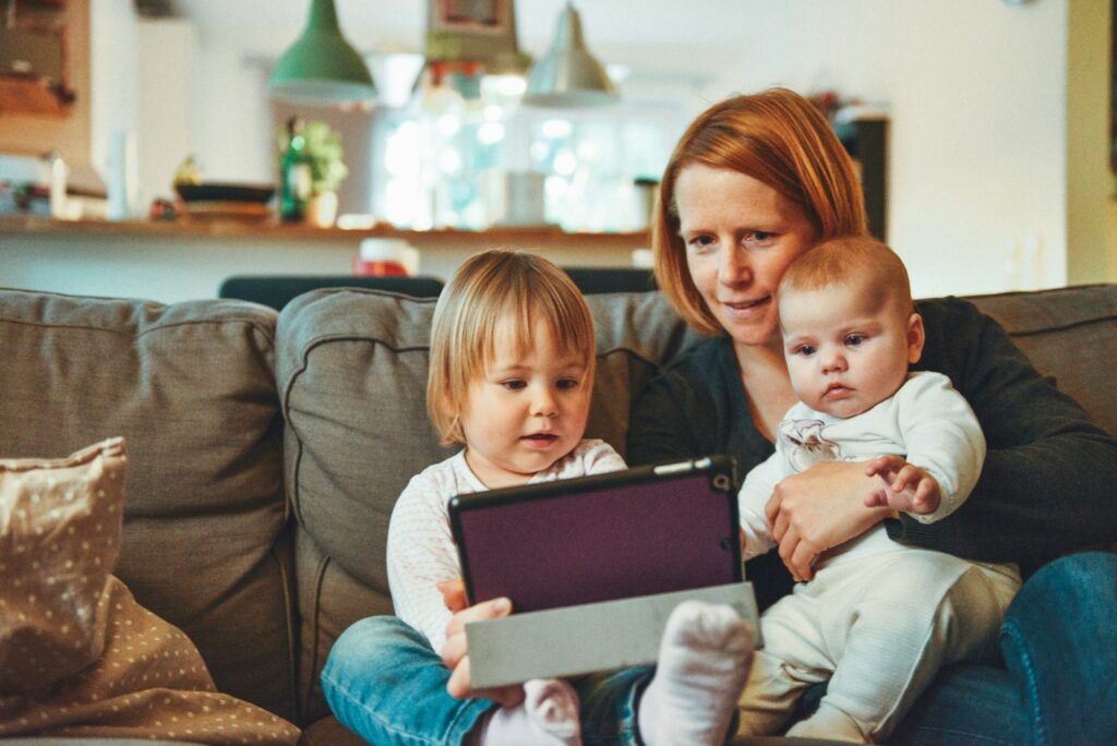 two babies and woman sitting on sofa while holding baby and watching on tablet representative of perinatal mental health
